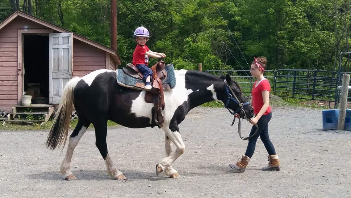 A young child on back of a black and white horse being led by a girl in red.